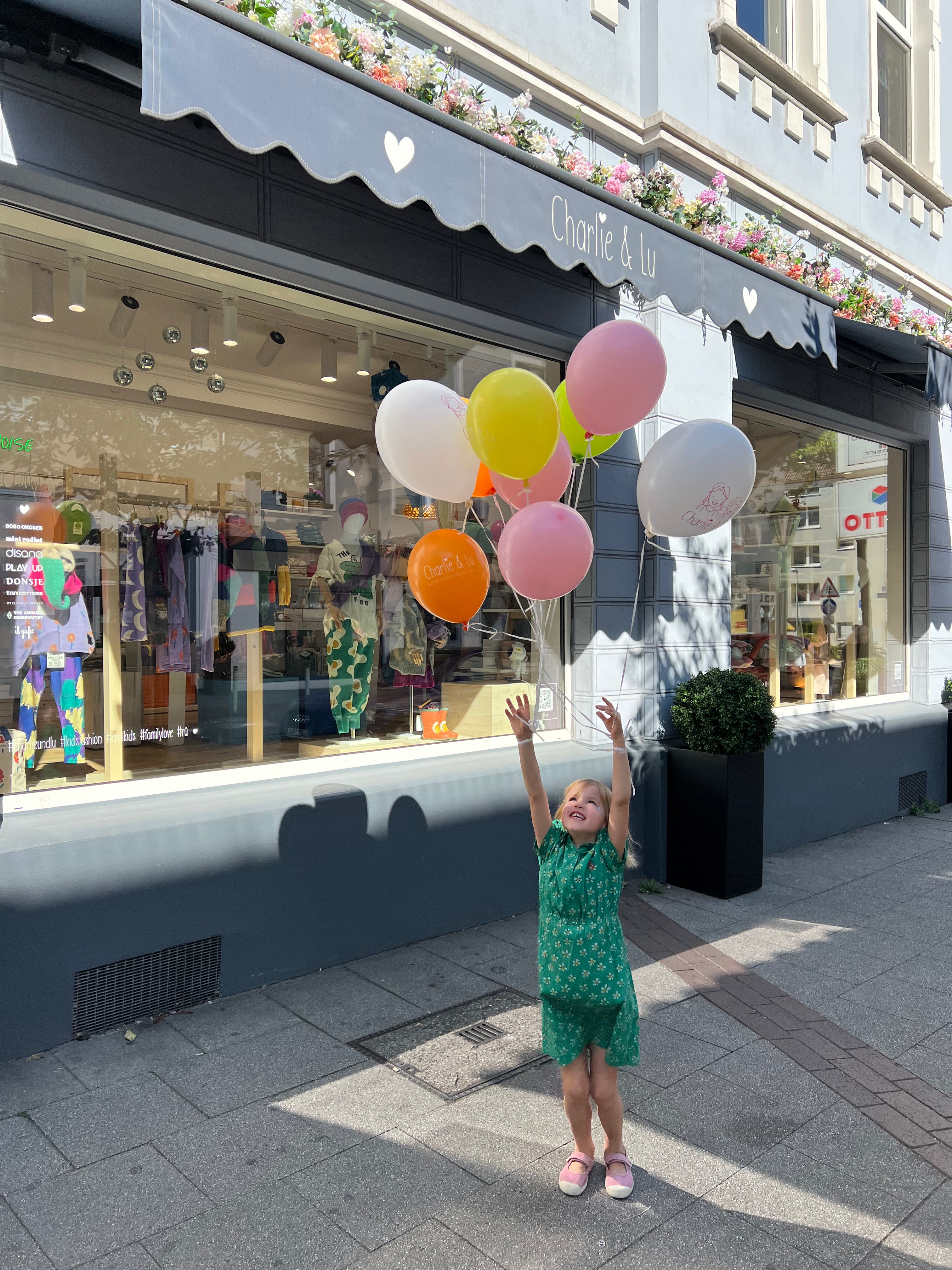 Außenansicht des Charlie & Lu Kids Fashion Stores in Essen Rüttenscheid, mit einem einladenden Schaufenster und einem kleinen Kind, das bunte Ballons steigen lässt. Die Szene unterstreicht die familienfreundliche und verspielte Atmosphäre des Stores.