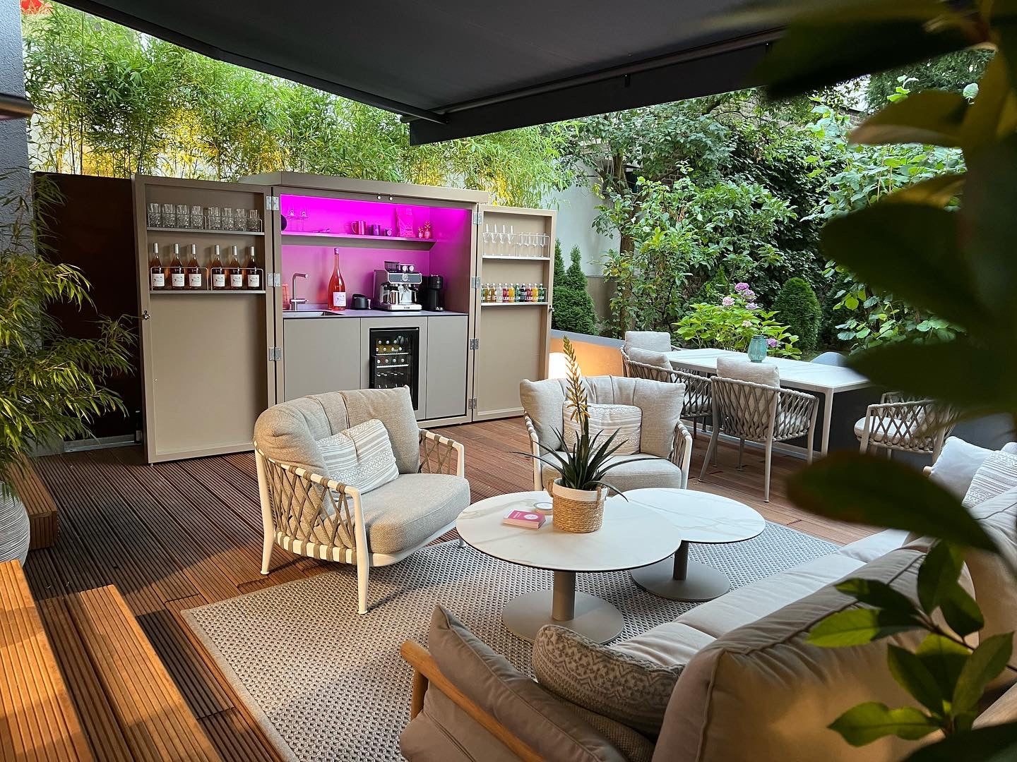 Gemütlicher Outdoor-Bereich mit modernen Loungemöbeln, einer integrierten Outdoor-Bar mit pinker Beleuchtung und umgeben von üppigem Grün. Der einladende Holzdeckboden und die stilvolle Einrichtung schaffen eine entspannte Atmosphäre für gesellige Momente